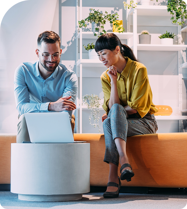 Deux collègues assis sur un canapé dans un bureau moderne et lumineux, souriant et regardant ensemble un ordinateur portable. L’une des personnes montre l’écran du doigt tandis que l’autre se penche avec attention, avec des étagères de plantes en arrière-plan.